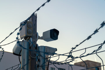 A camera is mounted on a pole next to a barbed wire fence. The camera is pointed at a building