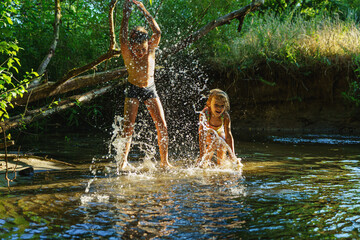 children playing in the river. A girl and boy raises her hands up in the water and splashes water drops - obrazy, fototapety, plakaty