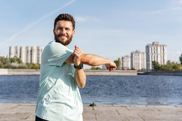 A man in casual sportswear stretches his arm by the waterfront, urban high-rises and clear skies...