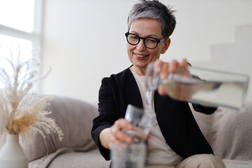 A senior woman enjoys a refreshing glass of water, embodying health and happiness in her home,...