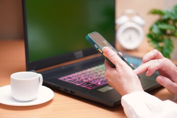 A smartphone is in the hands of a woman in a white blouse, working on a laptop with a green screen,...