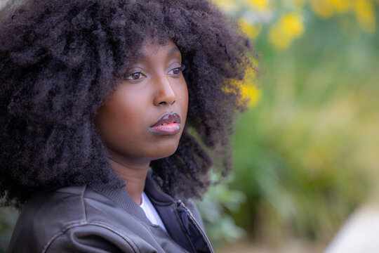 In a serene outdoor setting, a Black woman with a full afro hairstyle gazes into the distance. She is clad in a sleek leather jacket, embodying a blend of natures softness and urban edginess. The