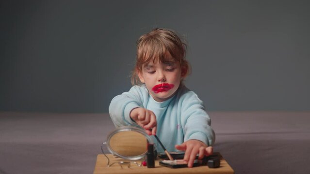 A cute little girl sitting in front of a mirror tries to do her makeup by dipping a brush into cosmetics