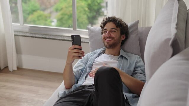 Satisfied caucasian guy is enjoying watching videos on couch. Carefree calm white man watch videos on phone, resting on sofa at home 
