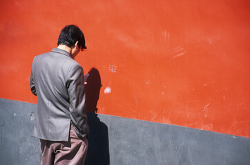 A man in a loose suit smoking a cigarette turned toward an orange wall looks at a flip phone. A boot print appears on the wall next to him.