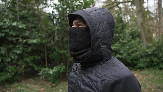 Young hispanic thug with mask and cap in black jacket standing on lookout.