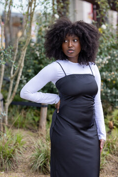 A Black woman stands confidently in an urban garden, her impressive afro hair accentuating her strong stance. She wears a white long-sleeved top under a black dress, blending casual style with an