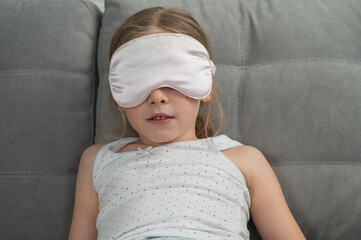 Little Caucasian girl puts on a sleep mask while sitting on the sofa. Doesn't want to sleep. 