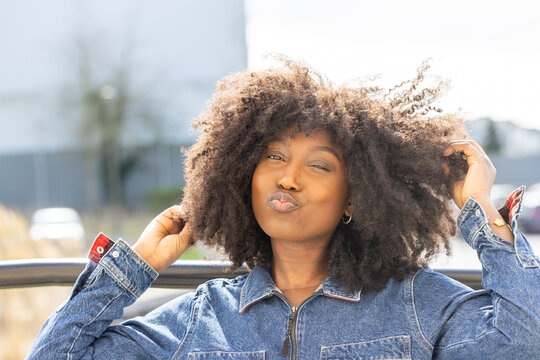 A light-hearted moment is captured as a Black woman with exuberant natural afro hair blows a kiss. She's donning a stylish denim outfit, sitting outdoors with a cityscape behind her. Her playful