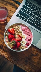 A vibrant smoothie bowl topped with fresh strawberries and granola next to a laptop, depicting a productive breakfast setting. - obrazy, fototapety, plakaty