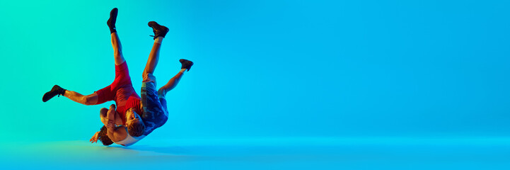 Two young male athletes in motion, wrestling, fighting against blue background in neon light....