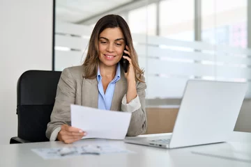 Poster Middle aged Hispanic professional business woman executive making call having conversation at work. Mature female manager or entrepreneur talking on the phone checking document sitting at office desk. © insta_photos