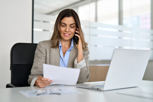 Fototapeta Middle aged Hispanic professional business woman executive making call having conversation at work. Mature female manager or entrepreneur talking on the phone checking document sitting at office desk.
