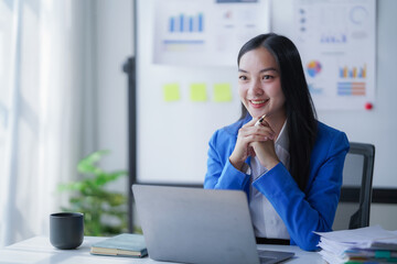 Smiling young Asian businesswoman in a modern office participating in an online video call, holding a pen and listening attentively, conveying professionalism, confidence and engagement in her role
