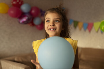 Smiling real girl 7 years old in yellow dress with blue balloon against festive backdrop in holiday at home - obrazy, fototapety, plakaty