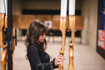 Focused young Asian woman choosing the right bow for her practice at an indoor archery range, depicting concentration and interest in the sport.