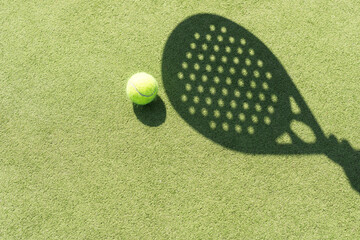 Shadow of a padel racket with a yellow ball on the green grass. - obrazy, fototapety, plakaty