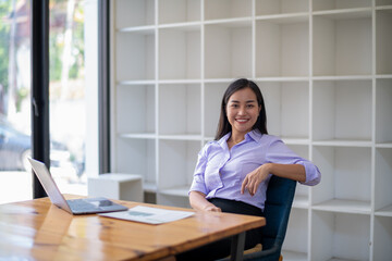 Smiling Asian businesswoman working at her desk with laptop, surrounded by minimalist style shelving in a contemporary office.