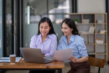 Two smiling businesswomen collaborating in a modern office environment, reviewing a document and using a laptop.
