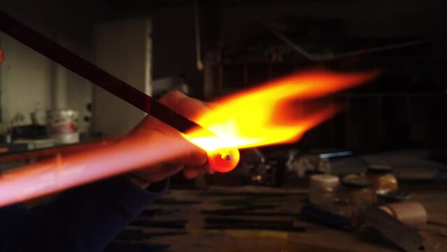 Close-up shot of hands shaping glass bead holding it over a gas torch in a workshop. Glass work jewelry