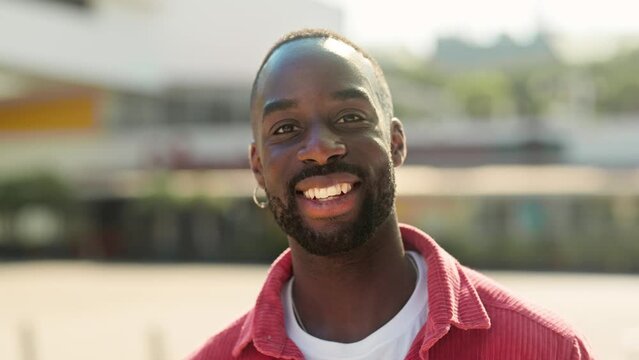 Smiling cool generation z Black hipster guy wearing red shirt posing outdoors. Close up portrait of happy young adult African man hipster standing on city street looking at camera.