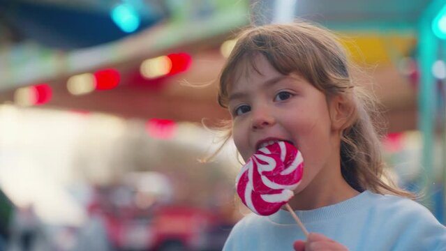 Little cute girl with a candy on a stick against the background of an amusement park shows different emotions of joy on her face