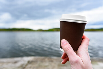 Paper coffee cup in hand by a lake.