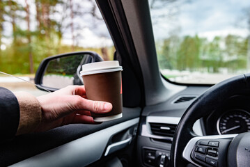 Driver holding paper cup with coffee at the car wheel.