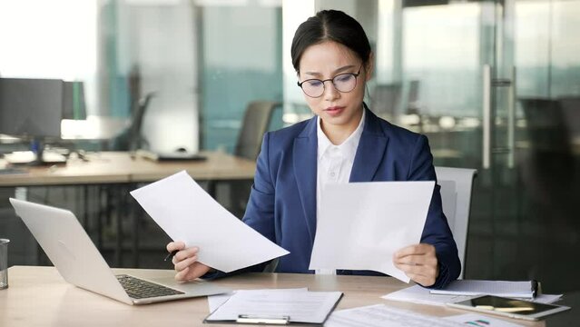 Confused young asian businesswoman having difficulty with paper work sitting at workplace in business office. Puzzled woman in formal suit looks at the documents and cannot understand the problem