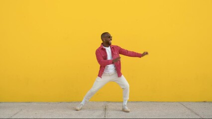 Happy excited cool stylish funky young African Black man dancer wearing sunglasses feeling joy having fun dancing, jumping standing at yellow color urban wall background. Full body.
