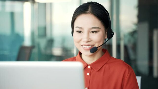 Asian woman in a headset customer service operator talking on a video call while sitting in business office. Call center agent advises the client by answering questions using laptop computer. Close up