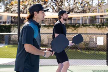 Two men playing pickleball together as a team. Each man is holding a paddle and waiting for the serve on an outdoor court. 