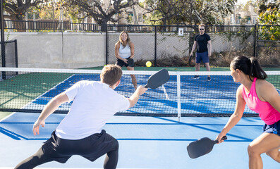 Two on two doubles pickleball game on an outdoor court. Four players are athletic and competitive hitting the ball over the next with their paddle. 