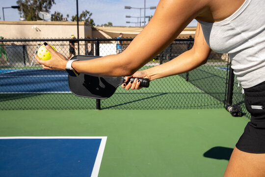 Detail shot of a female pickleball player preparing to serve. She is holding the yellow ball in one hand and her racket in the other during a game. 