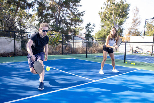 Pickleball players working on the court as a co-ed team. The man is hitting the pickle ball with his paddle. 