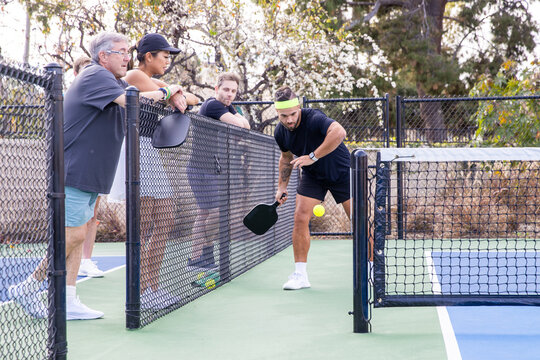 A pickleball player hits the pickle ball over a net on an outdoor court as other players watch and cheer. 