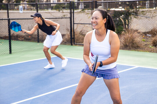 Two women playing a pickleball game as a team. The pair are smiling and having fun as they exercise on the outdoor court. 