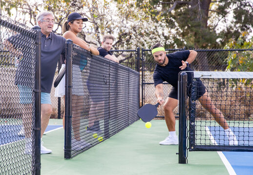 A pickleball player hits the pickle ball over a net on an outdoor court as other players watch and cheer. 