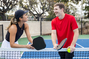 Pickleball coach giving a lesson to a student on an outdoor court. The Asian woman is teaching the Caucasian man with a mustache how to play the popular sport. 