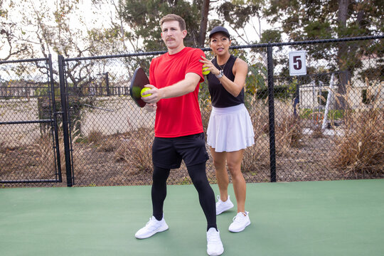 Pickleball coach giving a lesson to a student on an outdoor court. The Asian woman is teaching the Caucasian man with a mustache how to play the popular sport. 