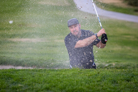 Golfer in the sand trap swinging to hit the golf ball on an uphill course. Sand is spraying onto the green. 