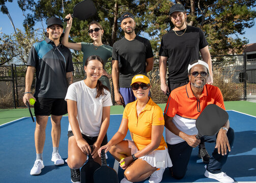 Group of seven friends together on pickleball court. The diverse players are laughing and smiling together outside and having fun. Multi-ethnic people and diversity in age and gender. 