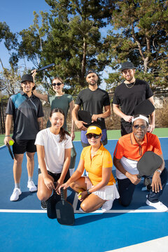 Group of seven friends together on pickleball court. The diverse players are laughing and smiling together outside and having fun. Multi-ethnic people and diversity in age and gender. 