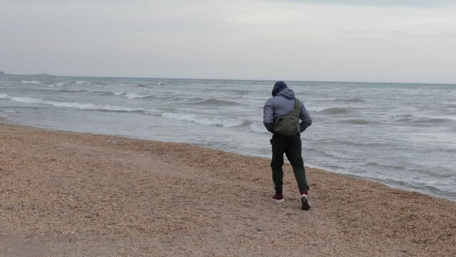 A walk in cool windy weather by the sea. A man walks along the beach in warm clothes. A beach with small shells on the shore of the Caspian Sea. Waves on the sea and strong wind.