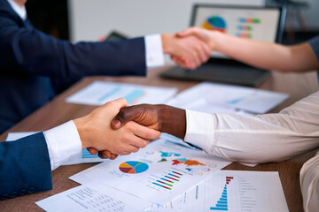 Multiracial business professionals seal agreement, shaking hands over data charts, teamwork,...