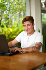 Transgender freelancer smiles at camera, works on laptop in a home office setting. Tattooed arms visible, light t-shirt, inclusive work environment vibe. Natural light, background of green plants.