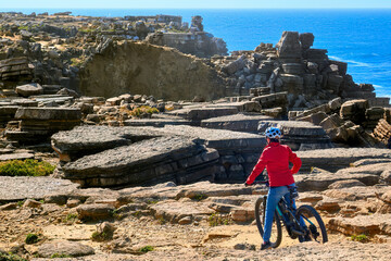 Brave senior woman riding her electric mountain bike on the rocky cliffs of Peniche at the western atlantic coast of Portugal - 789461209