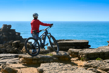 Brave senior woman riding her electric mountain bike on the rocky cliffs of Peniche at the western atlantic coast of Portugal - 789461261
