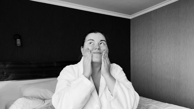 Black and white horizontal 4K video. Young adult white woman in bath robe applies moisturizer to face in hotel room. Concept of rest, skin protection during cold season, relax, body care, clean. Wife