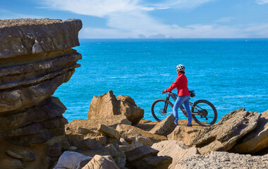 Brave senior woman riding her electric mountain bike on the rocky cliffs of Peniche at the western atlantic coast of Portugal - 789461491